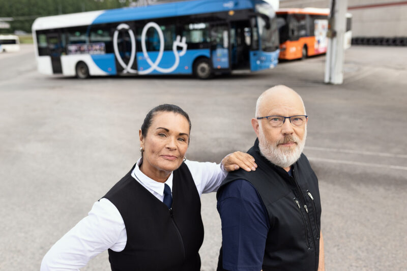 En busschaufför och en städare står framför några bussar som är parkerade på depån. Busschauffören håller sin hand på städarens axel.