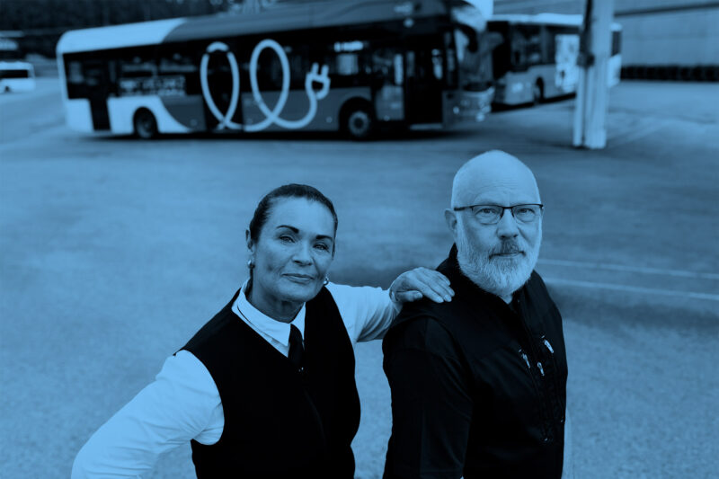 A bus driver and a cleaner stand in front of buses, and the bus driver has her hand on the cleaner's shoulder.