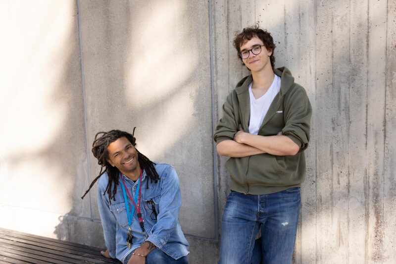 A youth worker is sitting and a construction painter is standing in front of a concrete wall.