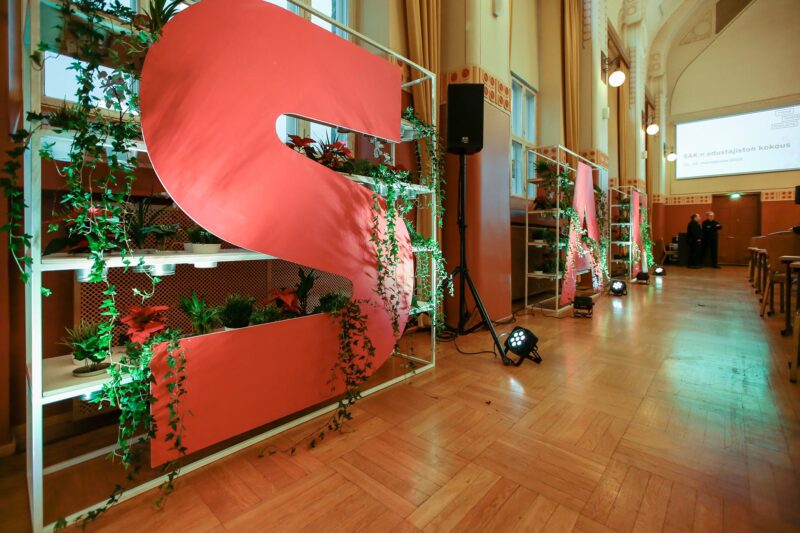 Large red letters S, A, and K attached to three shelves, which also have green plants placed on them.