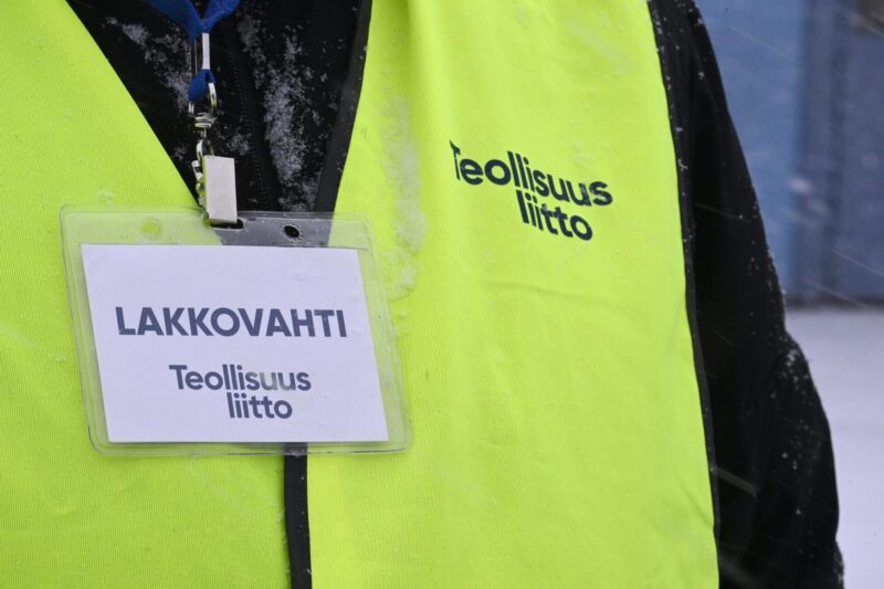 Close-up of a yellow safety vest worn by a strike guard, with a name tag attached that reads "Strike Guard ,Industrial Union" in Finnish.