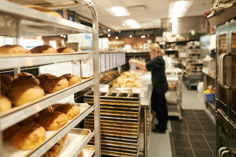 In the foreground, baked breads on baking trays; in the background, a bakery worker making breads.